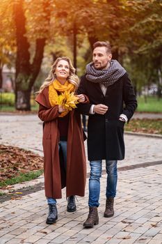 Walking arm-in-arm couple in the autumn park. Outdoor shot of a young charming couple in love walking along a path through a autumn park. Autumn toned image. Vertical image. 