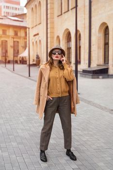 Full height self confident girl in an autumn beige coat and sunglasses, plaid panama hat standing posing on the street with coat on her shoulders. Retro toned photo. 