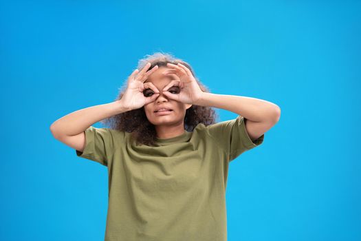 Funny young girl with funny pilot gesture African American woman looking positively at camera wearing peachy t-shirt showing her muscles isolated on blue background. Beauty concept. 