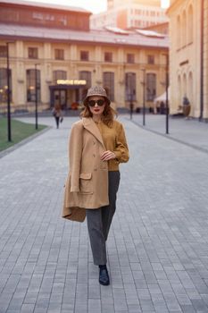 Confident fashion model young woman in sunglasses and hanging on a shoulder autumn beige coat walking on the street happily posing for the camera. Tinted photo. 