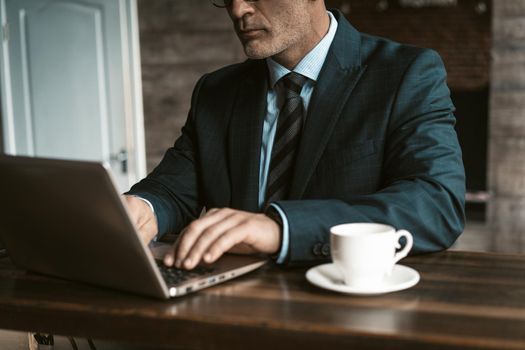 No face visible businessman sitting at the table working on laptop and drinking cup of coffee at the modern office. Man working on laptop in office. Business and finance concept.