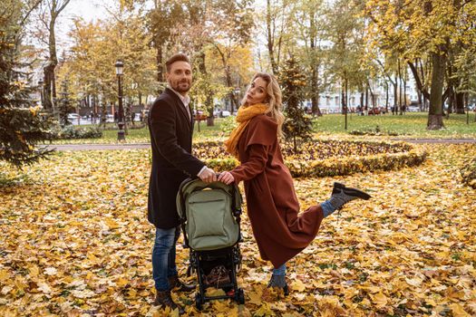 Happy family with a newborn baby in a stroller walking in an autumn park. Happy mother posing holding a stroller with a leg up outdoors in a golden autumn park. Tinted image. 