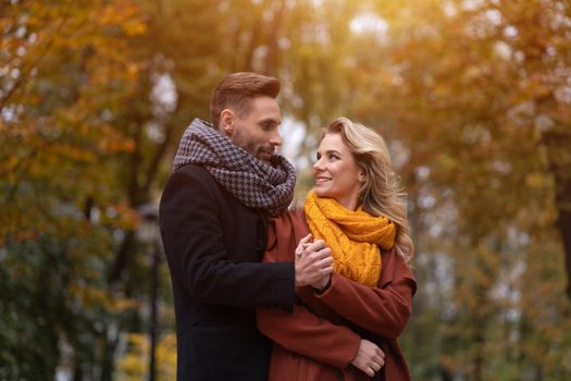 Portrait of a couple in love. Handsome man and a woman hugged from behind smile looking at each other in the autumn park. Outdoor shot of a young couple in love having great time. Autumn toned image. 