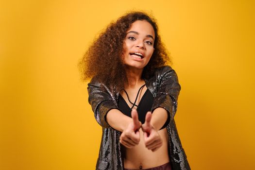 Hands front and thumbs up happy Afro American girl with beautiful hairstyle posing smile looking side away with hand on hips wearing shiny black jacket and black top on yellow background.
