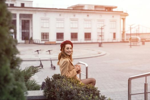 Beautiful french styled woman in an autumn coat and beige sits on a marbled flower beds with share it or rent electric scooters on background waiting for her date holding coffee mug. Toned photo. 