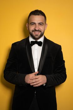 Smiling handsome man in black tuxedo with hands folded looking on camera, work profession lifestyle. Handsome young smiling caucasian man isolated on yellow background. Male fashion concept. 