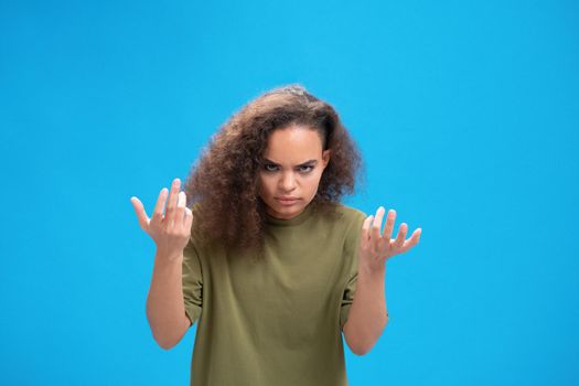 Unhappy african American millennial girl upset lifted hands up looking disappointedly at camera wearing olive t-shirt isolated on blue background. Human emotions, facial expression concept.