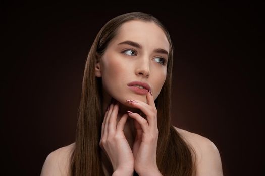 Beautiful spa model girl with perfect fresh clean skin and bare shoulders. Charming brunette model girl posing on camera touching her chin with a finger isolated on black background. 