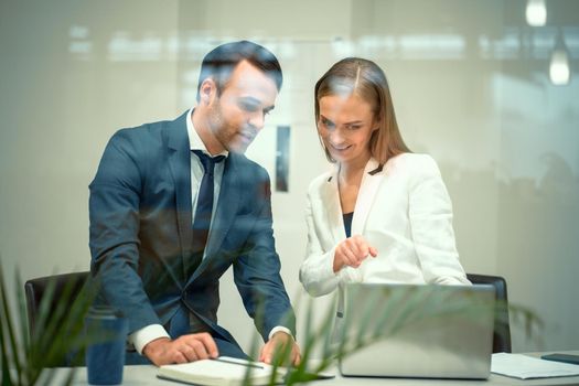 Handsome young businessman and a woman working on laptop in the office. Smiling diverse business partners young man and woman discussing project sitting in a modern bright meeting. 