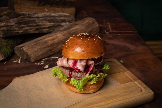 Fresh craft beef burger with fried slices of bacon and cherry sauce, fresh lettuce next to the decorative wooden lodges. Restaurant concept. Street food concept.