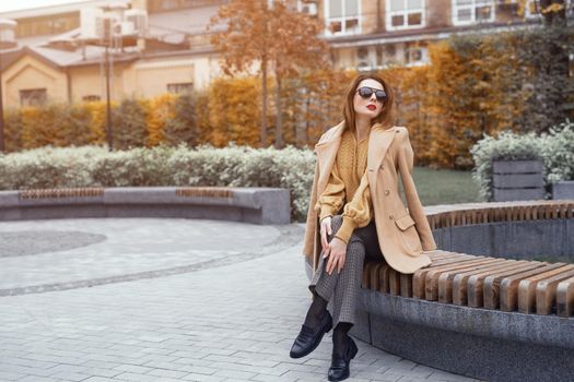 A beautiful girl in an autumn coat and beige sunglasses sits on a bench waiting for her date or girlfriends. Toned photo. 