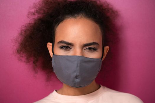 Questioning or studying look wearing reusable face mask African American charming girl in peachy t-shirt, to prevent others from corona COVID-19 and SARS cov 2 infection isolated on pink background. 