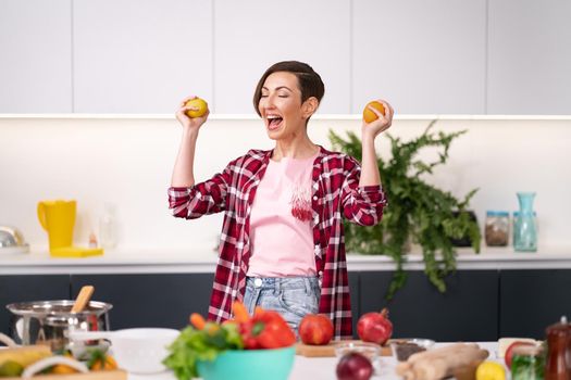 Holding an lemon and orange in hands happy woman dancing at kitchen. Charming housewife wearing plaid shirt with a short hair while standing at the kitchen of a new house. 