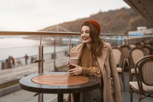 Happy young girl sitting outdoors street restaurant or terrace looking smiling away holding a coffee mug. Portrait of stylish young woman wearing autumn coat and red beret outdoors. 