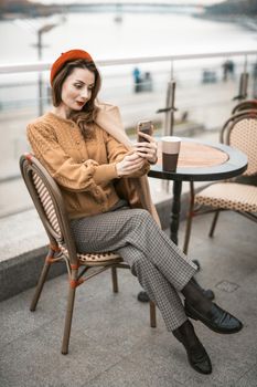 Charming french young woman using her smartphone taking selfie while having cup of coffee. Frenchwoman wearing red beret sitting on terrace of restaurant or cafe with background of autumn urban city. 