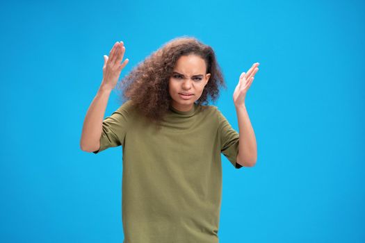 Angry african American girl lifted her hands up looking disappointedly at camera wearing olive t-shirt isolated on blue background. Human emotions, facial expression concept.