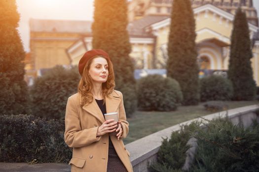 Pensive beautiful young woman holding a cup of coffee on the street female fashion. Portrait of stylish young woman wearing autumn coat and red beret outdoors. Autumn accessories. 