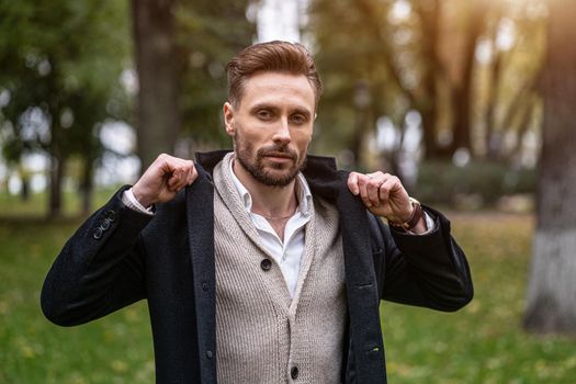 Handsome man holing a corners of his trench coat standing outdoors in an autumn park smiling looking at the camera. Handsome young man in dark blue coat and scarf or muffler. 