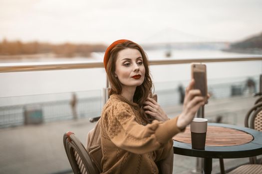 Beautiful young woman takes selfie using her smartphone while having cup of coffee. Frenchwoman wearing red beret sitting on terrace of restaurant or cafe with background of autumn urban city. 