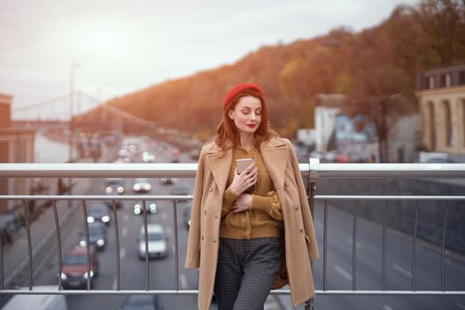 Portrait of stylish young woman wearing autumn coat and red beret outdoors. Tender young woman warming up with cup of coffee on the street female fashion standing on a pedestrian bridge. Toned image. 