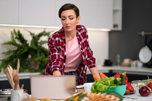 Young woman cooking in the kitchen. Using a laptop computer while cooking young housewife searching online recipes or watching online tv show while cooking. Healthy life living. 