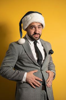 Overeating, improper nutrition during the New Year holidays handsome bearded man in Santa blue hat and grey suit, looking at camera isolated on yellow background. 