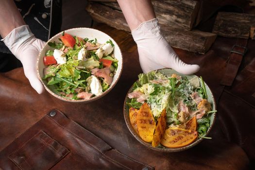 Two bowls of vegetable salad with salmon, mozzarella cheese and grilled slices of fruits stands on a table covered with a leather rag. Restaurant concept. Vertical image.