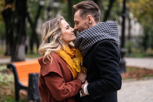 Passionate couple handsome guy hold his girlfriends hands and touching noses stand in fall town park wearing coats and scarfs. Smiling couple hugging in autumn park. 