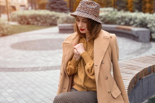 Parisian girl in beige coat and plaid panama hat siting on rounded bench waiting for her date or girlfriends. Autumn walk concept. Retro tinted photo.