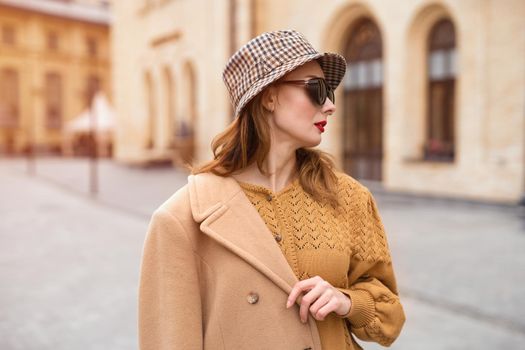 Charming model girl in an autumn beige coat and sunglasses, plaid panama hat standing with face turned holding coat on her shoulders. Retro toned photo. 