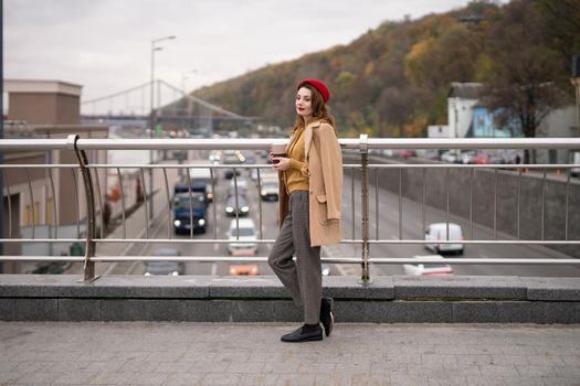 Charming french young woman drinks coffee using a coffee mug looking at camera smiling standing at pedestrian bridge with cars on the road. Looking happy fashion girl in a red beret and beige coat. 