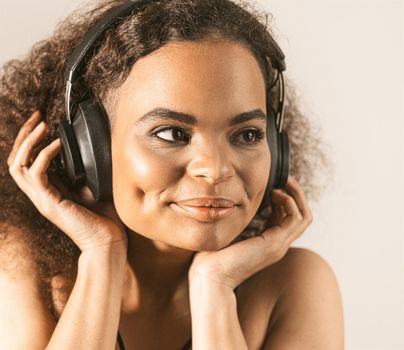 Young African-American girl listening music in headphones wearing black top isolated on grey background, emotionally move. Concept of emotions, facial expression. Square cropped. 