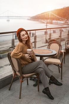 Charming woman enjoying her cup of coffee sitting in a spring outdoor cafe. French woman in red beret with background of urban city. Parisian woman with coffee mug on city street in evening. 