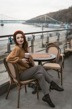 Sitting sideways turned Parisian young woman in restaurant terrace. Portrait of stylish young woman wearing autumn coat and red beret outdoors. 