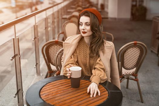 All thoughtful in memories young Parisian woman sitting outdoors restaurant terrace looking away holding a coffee mug. Portrait of stylish young woman wearing autumn coat and red beret outdoors. 