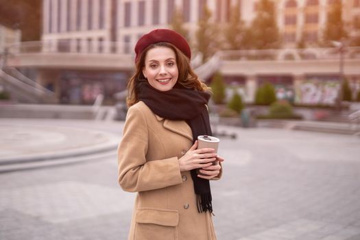 Parisian young woman holding reusable mug with coffee standing outdoors. Portrait of stylish young woman wearing autumn coat and red beret outdoors. Autumn accessories. 