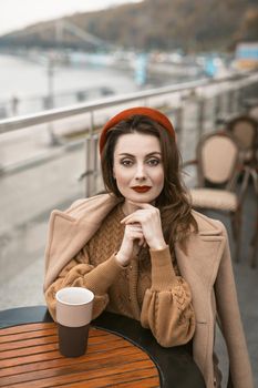 Beautiful french woman drink coffee using coffee mug sitting at restaurant terrace with coffee mug looking at camera. Portrait of stylish young woman wearing autumn coat and red beret outdoors. 