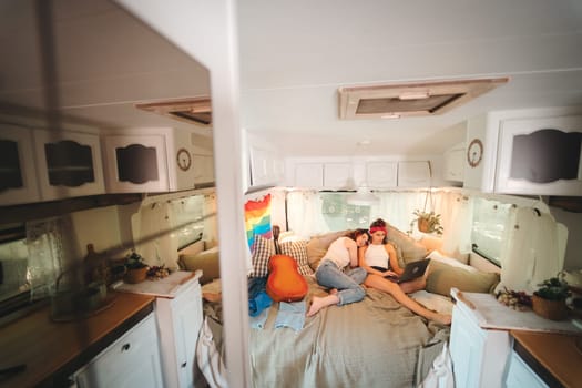 Portrait of a cute lesbian couple. Two girls spend time tenderly together watching movie on laptop in a camper trailer with LGBT flag on the wall. Love and attitude. LGBT concept. High quality photo