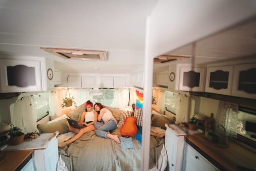 Portrait of a cute lesbian couple. Two girls spend time tenderly together watching movie on laptop in a camper trailer with LGBT flag on the wall. Love and attitude. LGBT concept. High quality photo