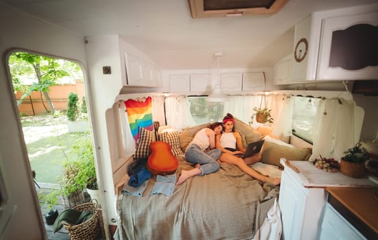 Portrait of a cute lesbian couple. Two girls spend time tenderly together watching movie on laptop in a camper trailer with LGBT flag on the wall. Love and attitude. LGBT concept. High quality photo