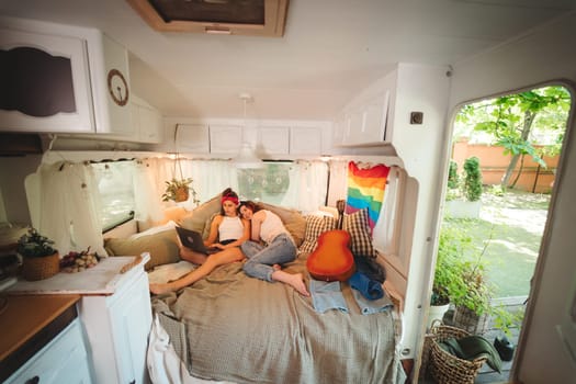 Portrait of a cute lesbian couple. Two girls spend time tenderly together watching movie on laptop in a camper trailer with LGBT flag on the wall. Love and attitude. LGBT concept. High quality photo