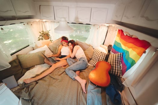 Portrait of a cute lesbian couple. Two girls spend time tenderly together watching movie on laptop in a camper trailer with LGBT flag on the wall. Love and attitude. LGBT concept. High quality photo