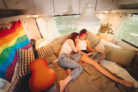 Portrait of a cute lesbian couple. Two girls spend time tenderly together watching movie on laptop in a camper trailer with LGBT flag on the wall. Love and attitude. LGBT concept. High quality photo