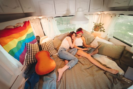 Portrait of a cute lesbian couple. Two girls spend time tenderly together watching movie on laptop in a camper trailer with LGBT flag on the wall. Love and attitude. LGBT concept. High quality photo