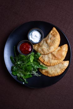 top view fried chebureks with sauce and herbs on a black plate.