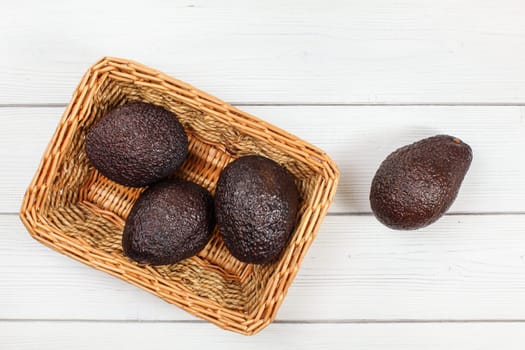 Flat lay photo - dark brown avocados ( bilse variety ) in square wooden basket on white boards desk.