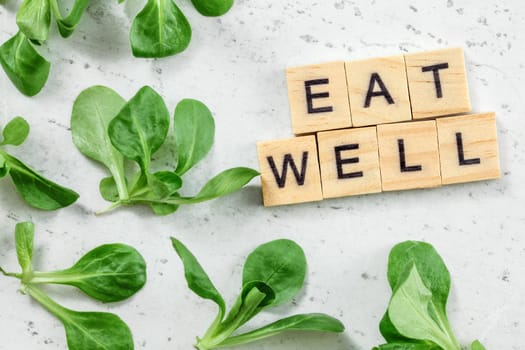 Flat lay photo - leaves of cornsalad ( Valerianella locusta ) with text EAT WELL at wooden blocks on white working board. Green leaves healthy eating concept.