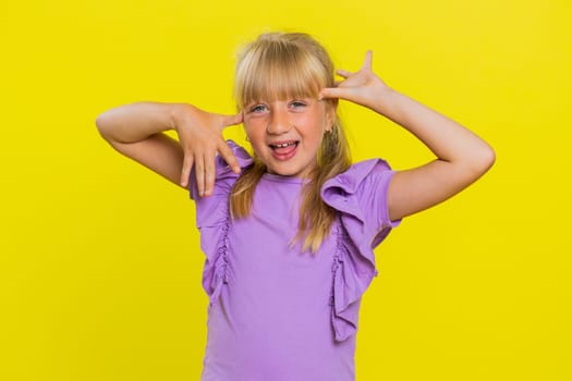 Cheerful funny bully young cute girl showing tongue making faces at camera, fooling around, joking, aping with silly face, teasing. Preteen blonde child kid on yellow background. Little children