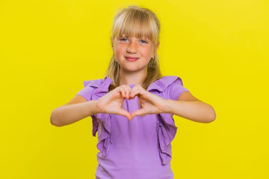 I love you. Smiling young cute blonde school girl makes heart gesture demonstrates love sign expresses good feelings and sympathy. Happy preteen female child kid isolated on studio yellow background