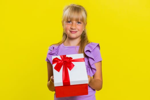 Lovely smiling young preteen child girl kid presenting birthday gift box stretches out hands, offer wrapped present career bonus, celebrating party. Little cute children on studio yellow background